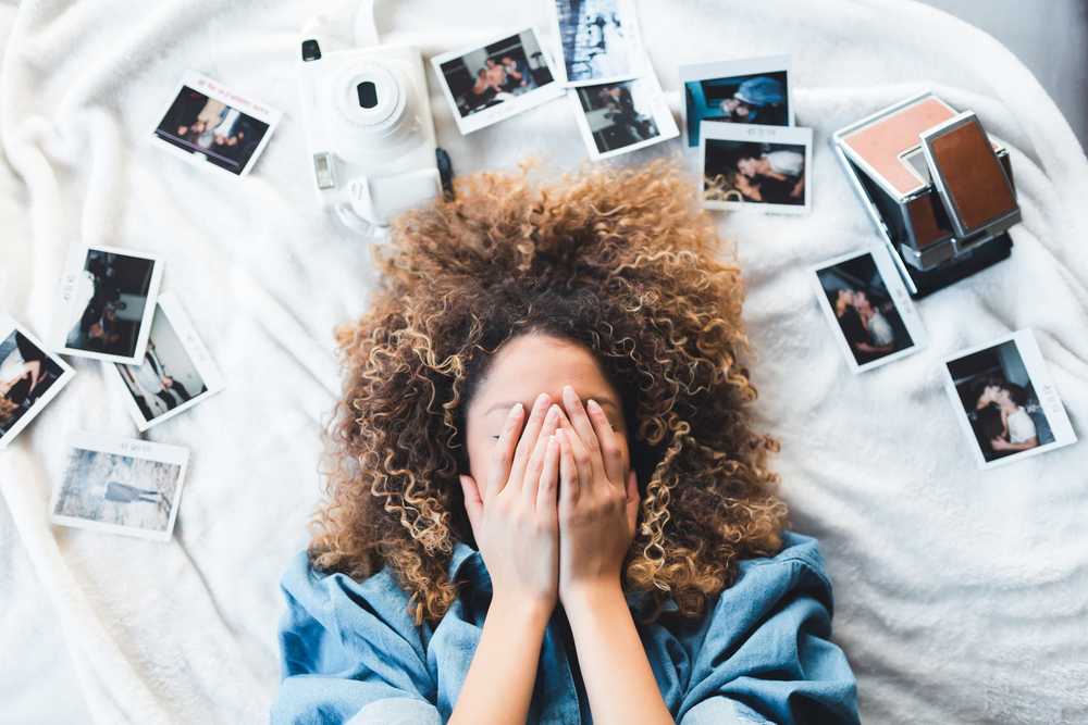 Woman lying on bed covering her face surrounded by photos and white camera